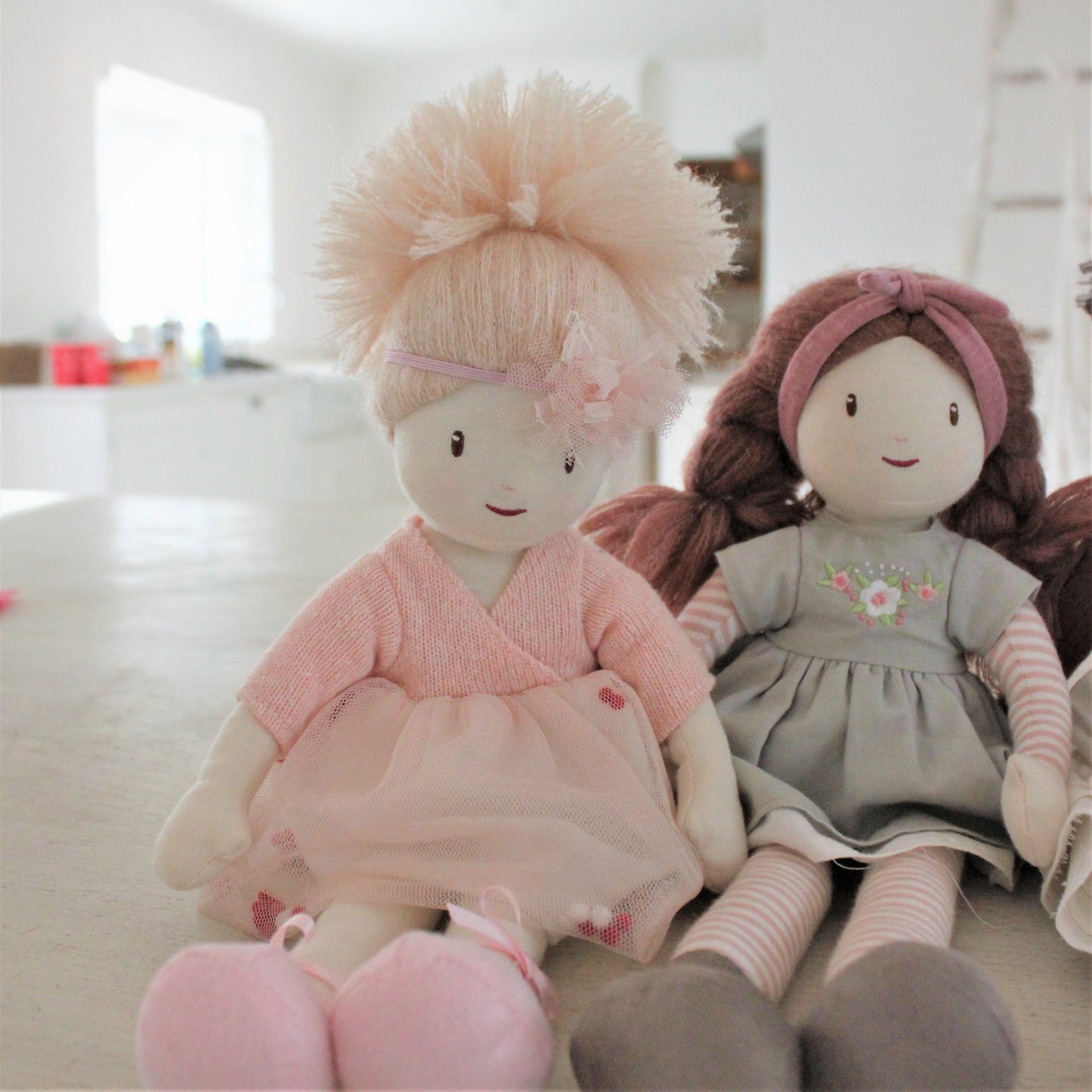 Two dolls, one in pink outfit and the other in gray dress with floral details, sitting on a light-colored surface.
