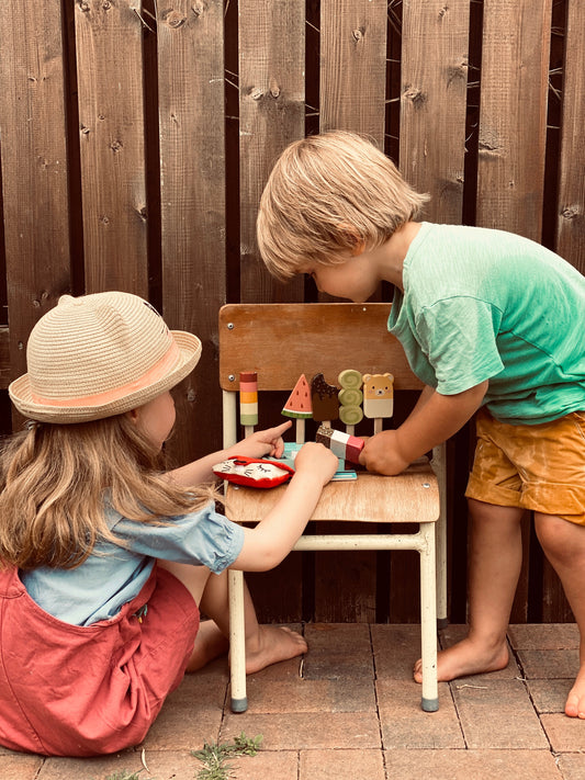 Two children playing with a small wooden table outdoors against a wooden fence.