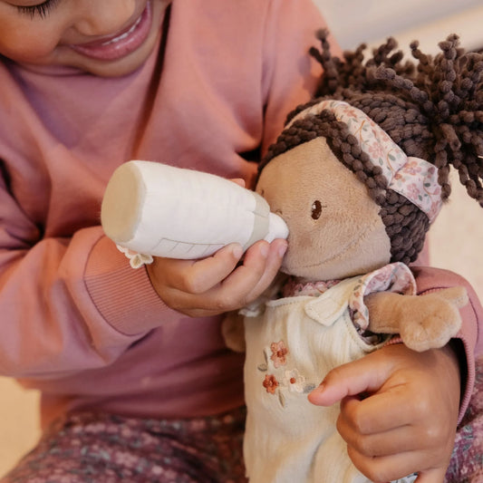 Child holding a bottle and a doll with braided hair, wearing a pink outfit.