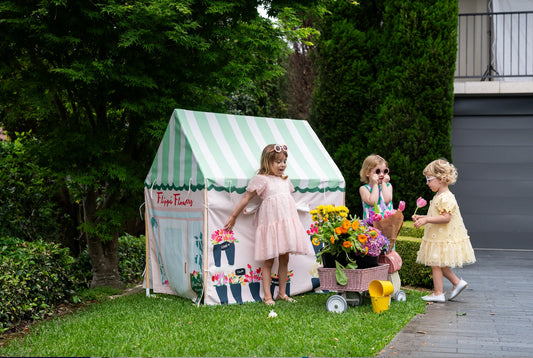 Three young girls play outdoors on a grassy lawn next to a small green and white striped playhouse tent. One girl in a pink dress stands in front of the tent, while the other two girls in pastel dresses stand beside a small cart filled with colorful flowers and watering cans.