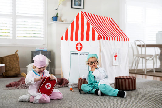 Two children sitting on a carpeted floor in a playroom, dressed as doctors with a red and white striped play tent, a red first-aid kit, and toy medical accessories.