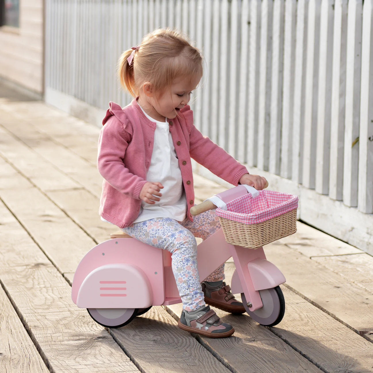 Child on a pink toy scooter with a basket on a wooden deck