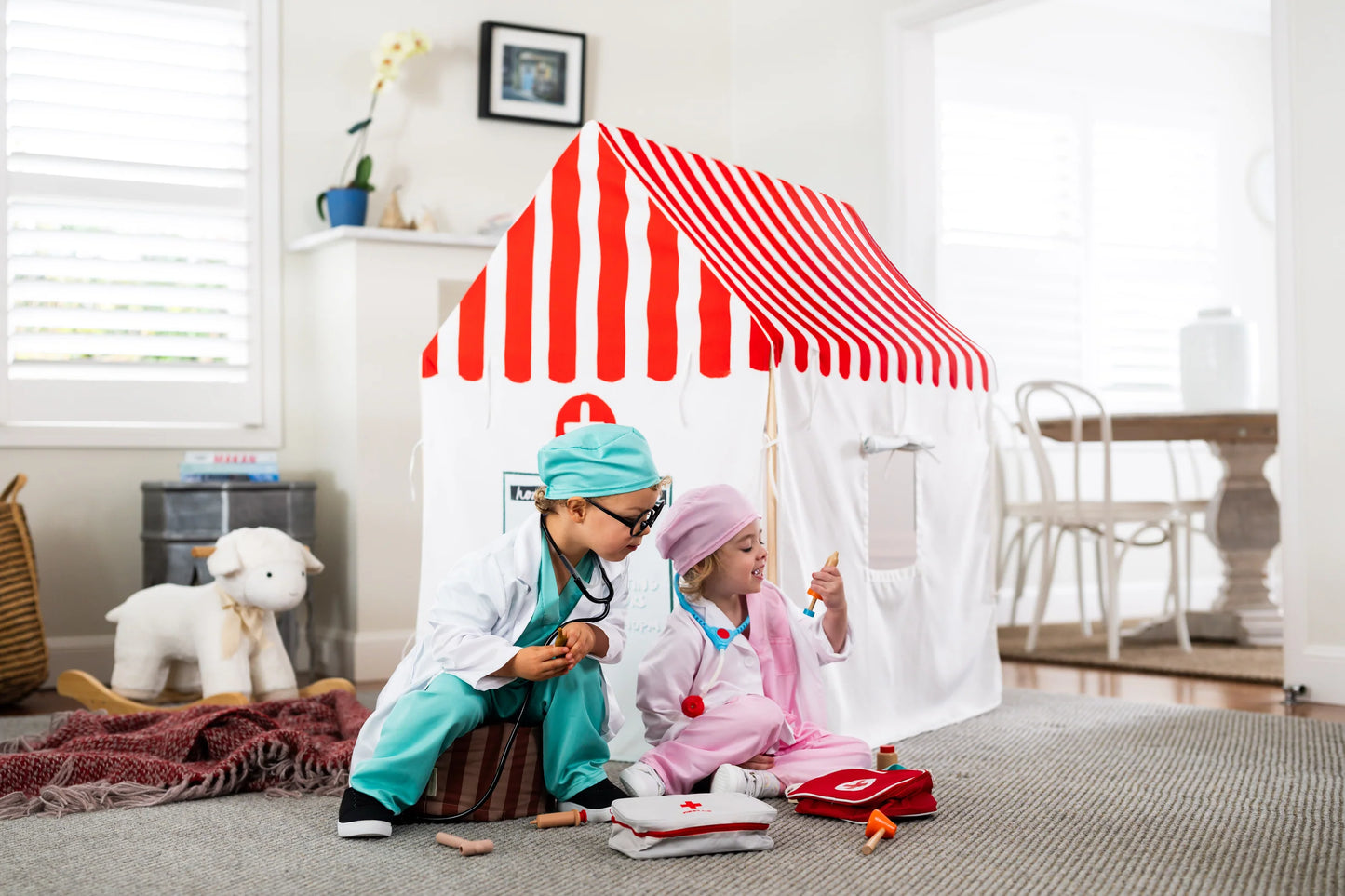 Two children dressed as doctors play with a toy stethoscope and medical playset in a bright playroom. One child wears a white lab coat and teal scrubs while the other wears a pink outfit, positioned in front of a small white and red striped doctor playhouse tent.