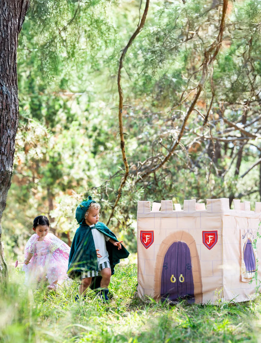 A children's play castle made from canvas with a printed medieval theme, featuring a purple door and red shield emblems, set in a grassy wooded outdoor play area with two children playing nearby.