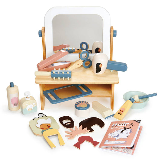 Children's toy hair salon set with wooden table, accessories including brush, hairdryer, straighteners, scissors, and hair styling products, and a magazine.
