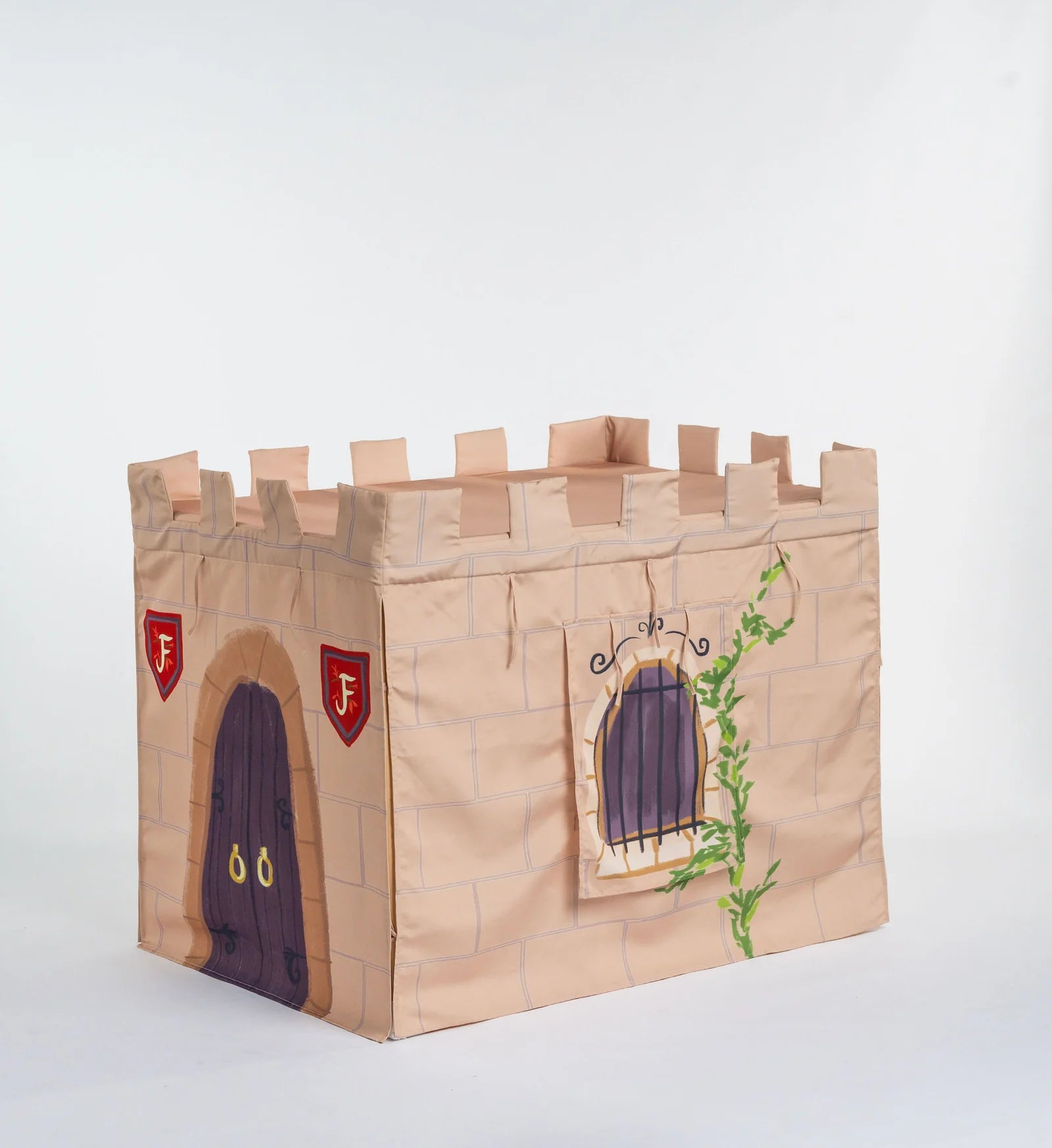 A children's play castle toy made from tan canvas or fabric, printed with a stone wall pattern, a purple door, red shield emblems with white letters, and a green vine climbing a window.