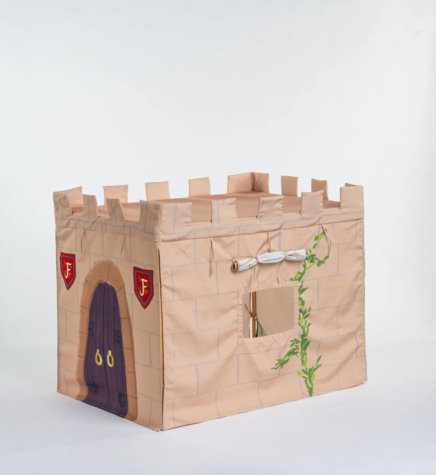 A cardboard castle playhouse with crenellated top, a purple door, red shields with gold letters, and a green vine decoration, photographed on a light gray studio background.