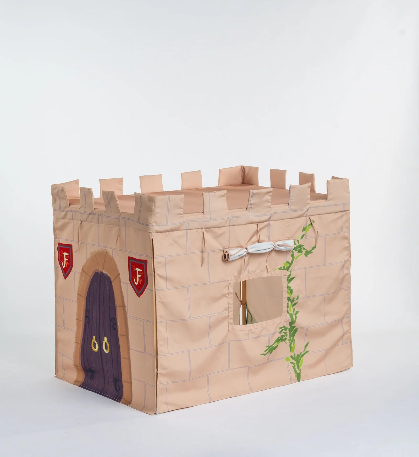 A cardboard castle playhouse with crenellated top, a purple door, red shields with gold letters, and a green vine decoration, photographed on a light gray studio background.