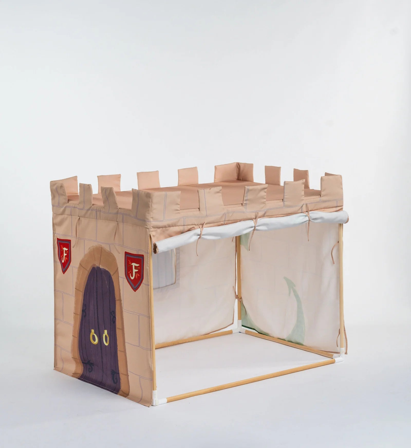 A children's play castle tent made from cardboard and fabric, featuring a tan brick-patterned front wall with a dark purple door, red shield-shaped badges with white letters, and beige canvas side panels, photographed on a light gray studio background.