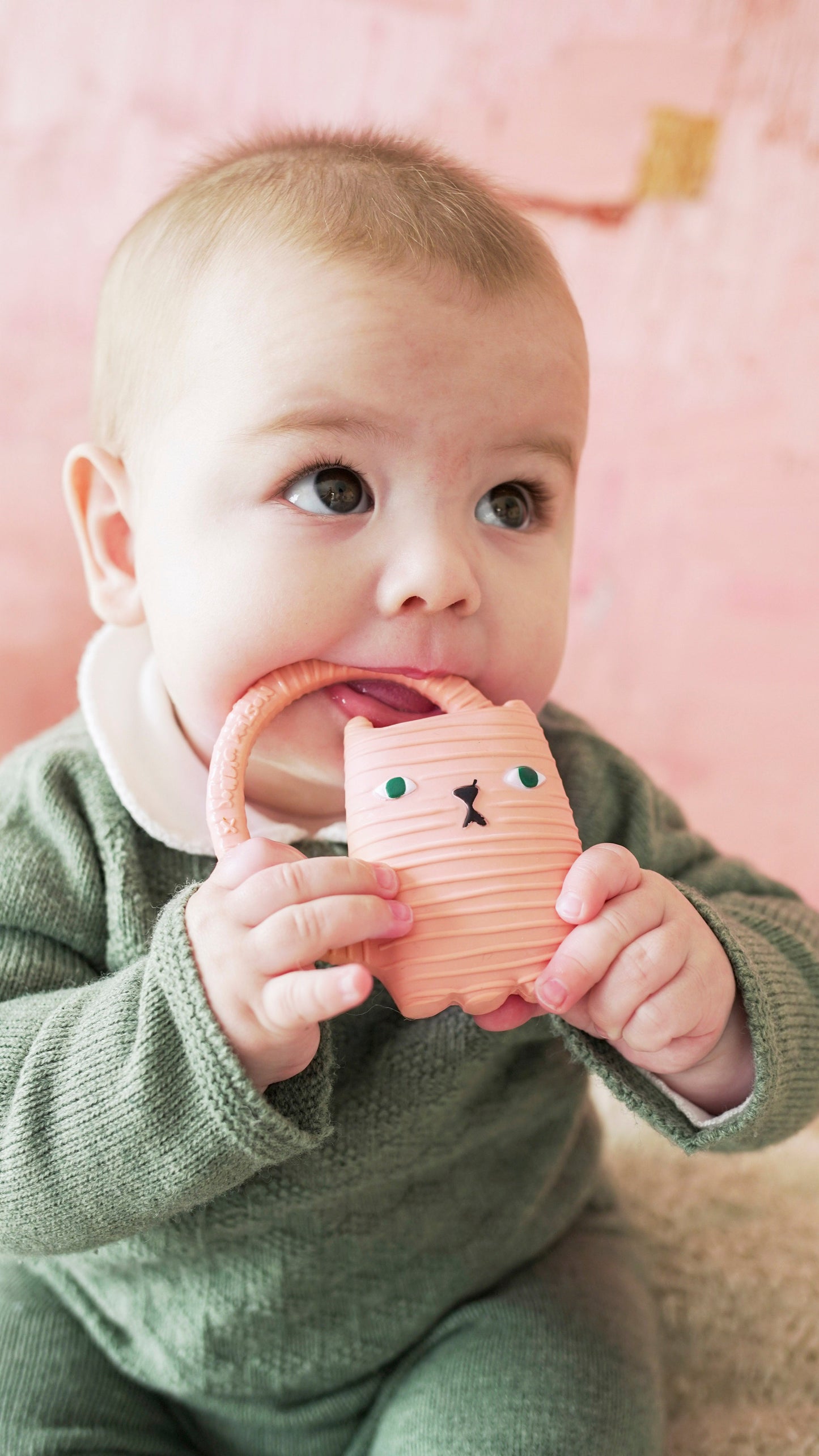 Baby holding a pink silicone teether with a bear design against a pink background