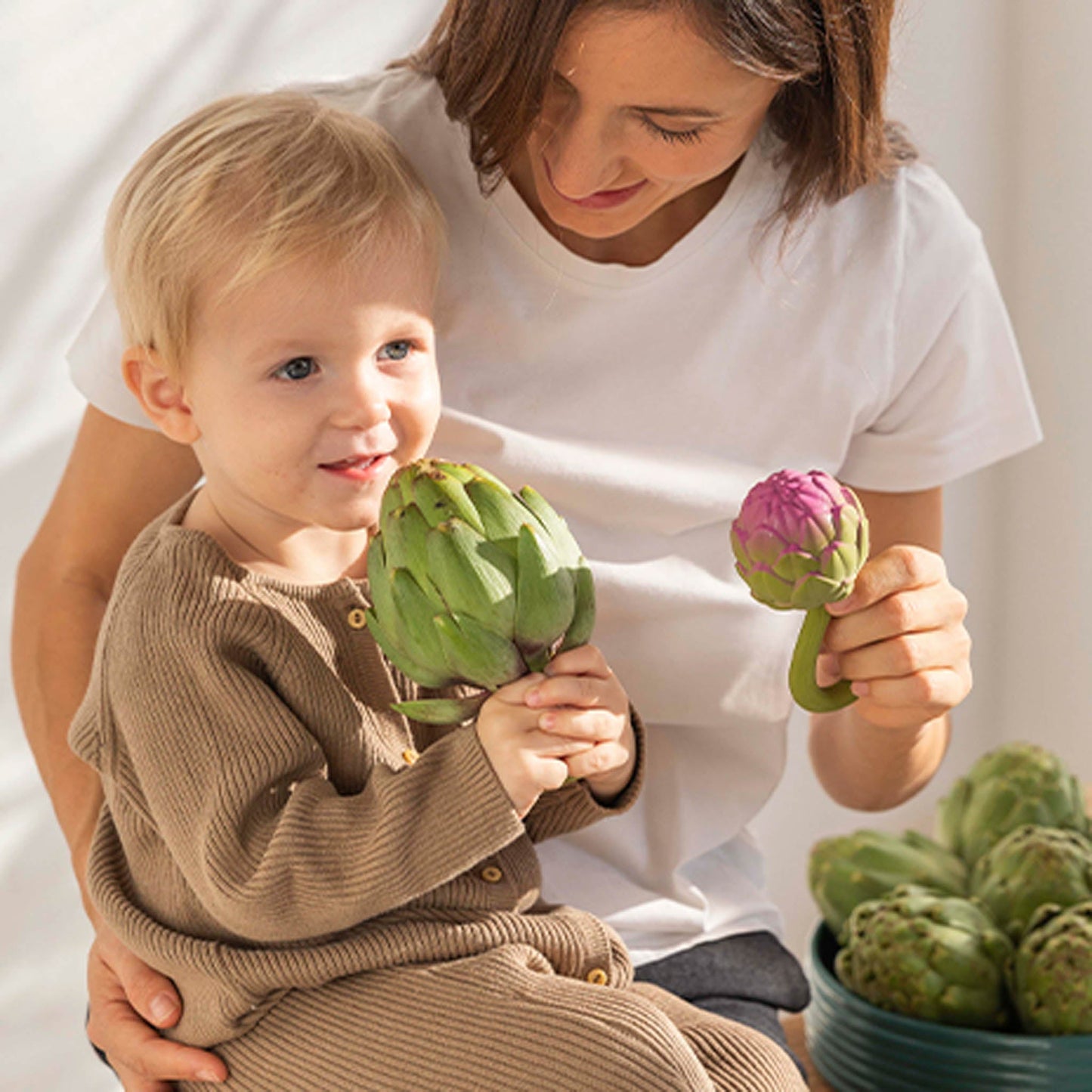 Woman and child holding artichokes together with a bowl of artichokes in the background.