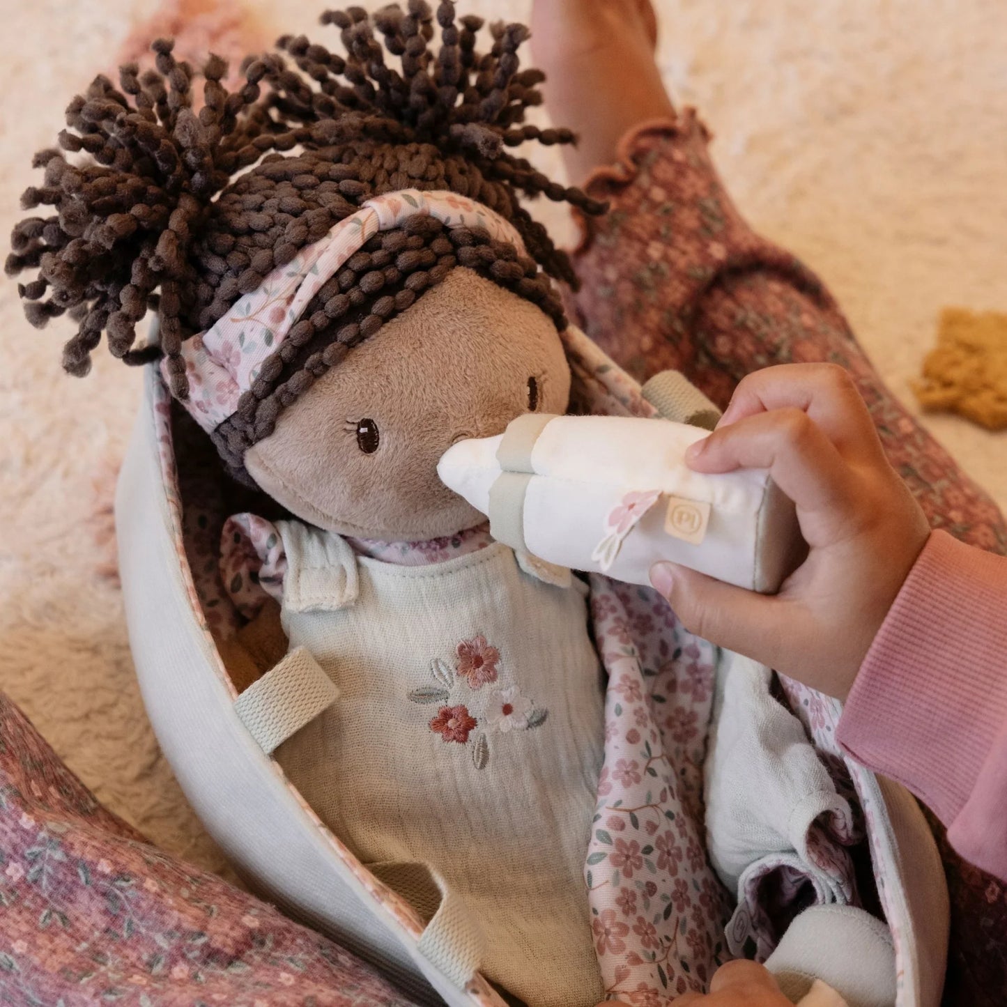 Person holding a bottle to a doll with braided hair, wearing a floral outfit.