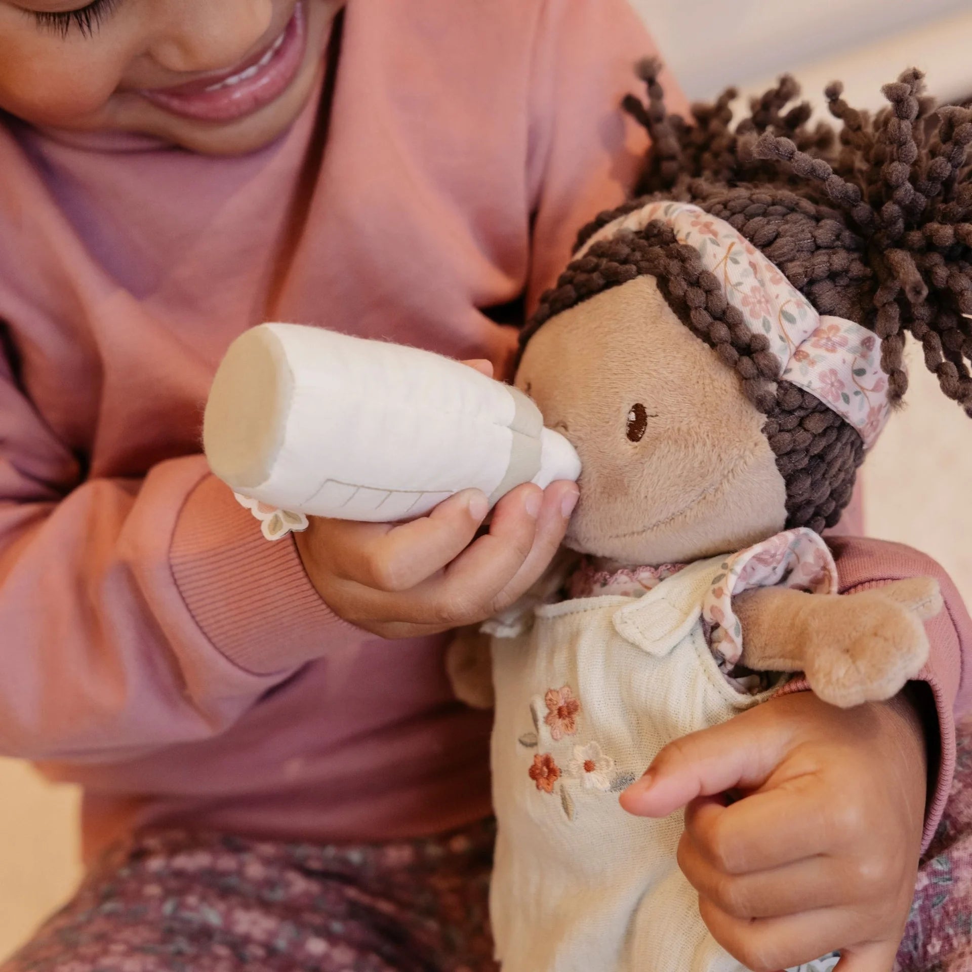Child holding a bottle and a doll with braided hair, wearing a pink outfit.