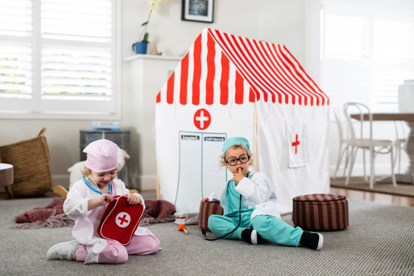 Two children sitting on a carpeted floor in a playroom, dressed as doctors with a red and white striped play tent, a red first-aid kit, and toy medical accessories.