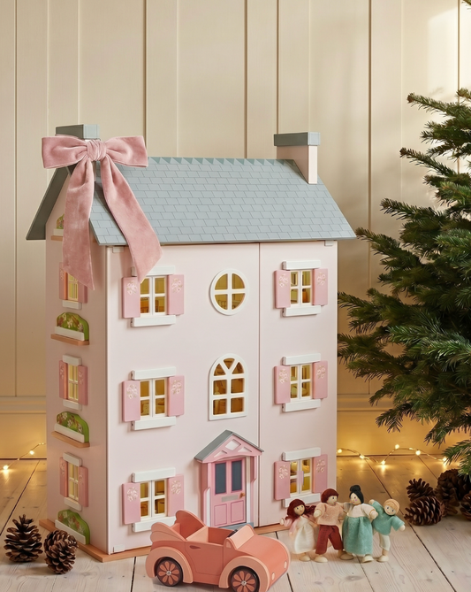 A pastel pink dollhouse-style house with a gray roof, pink shutters, and a large pink bow on top, styled on a wooden floor with a small pink car, miniature figures, pinecones, and a Christmas tree in the background.