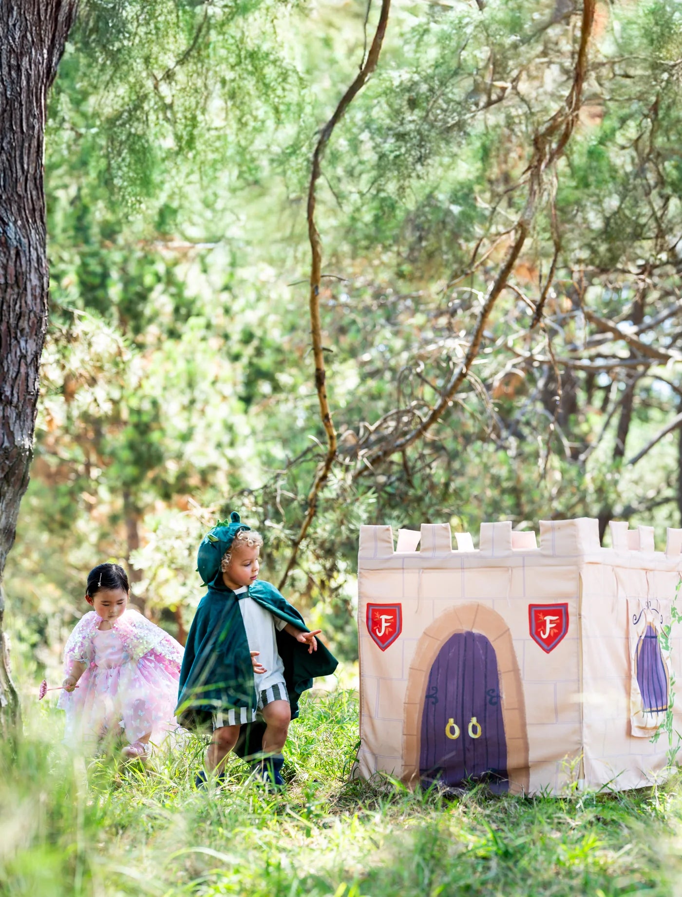 A children's play castle made from canvas with a printed medieval theme, featuring a purple door and red shield emblems, set in a grassy wooded outdoor play area with two children playing nearby.