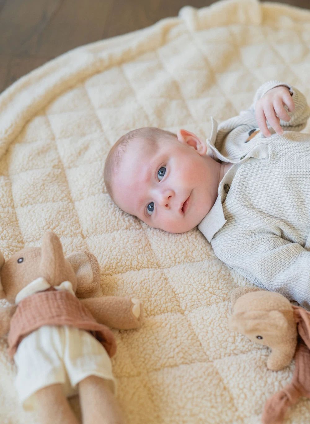 A baby lying on a cream-colored quilted blanket with plush stuffed animals nearby.