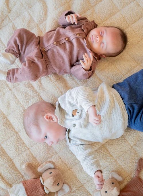 Two babies lying on a beige quilted blanket, wearing matching knit outfits. One baby wears a dusty mauve button-front romper, and the other wears a cream knit top and blue pants.