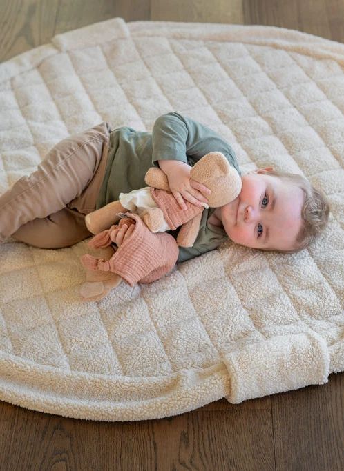A baby lying on a large cream quilted blanket on a wooden floor, holding a plush toy.