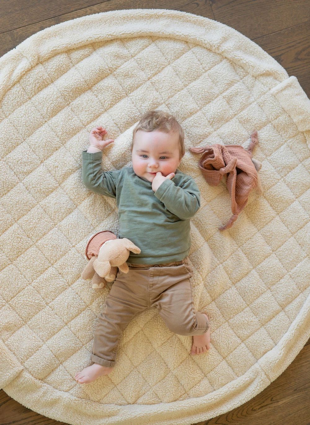 A baby lying on a round quilted cream play mat on a wooden floor, wearing a sage green long-sleeve top and tan pants, with a small stuffed bunny and a dusty pink cloth toy nearby.