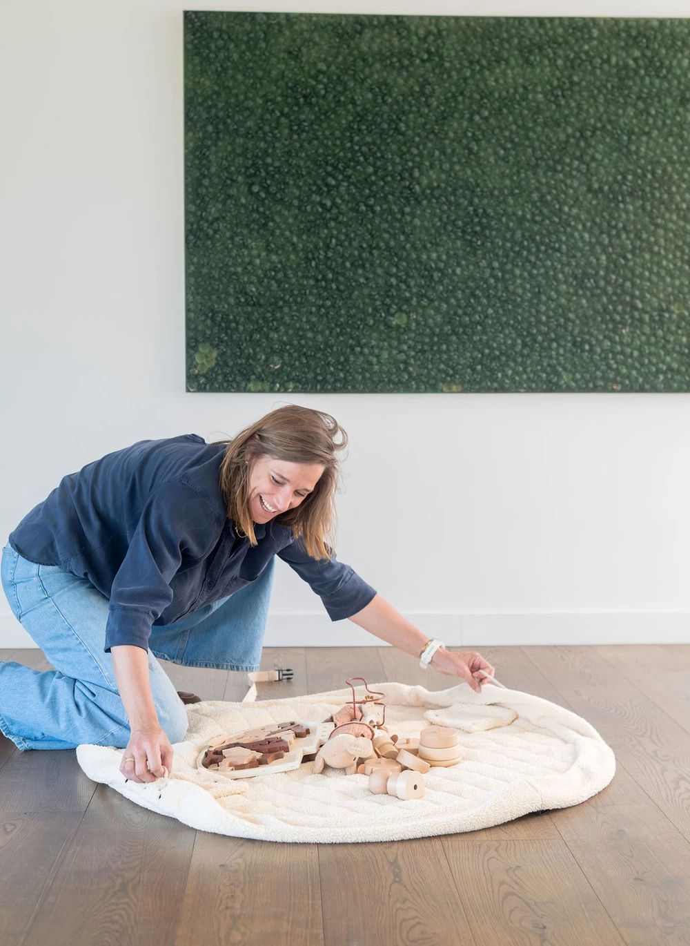 A woman kneels on a wooden floor in a bright room, arranging small wooden beads and a small bag on a round cream-colored blanket or rug. A large green textured wall hanging is visible behind her.