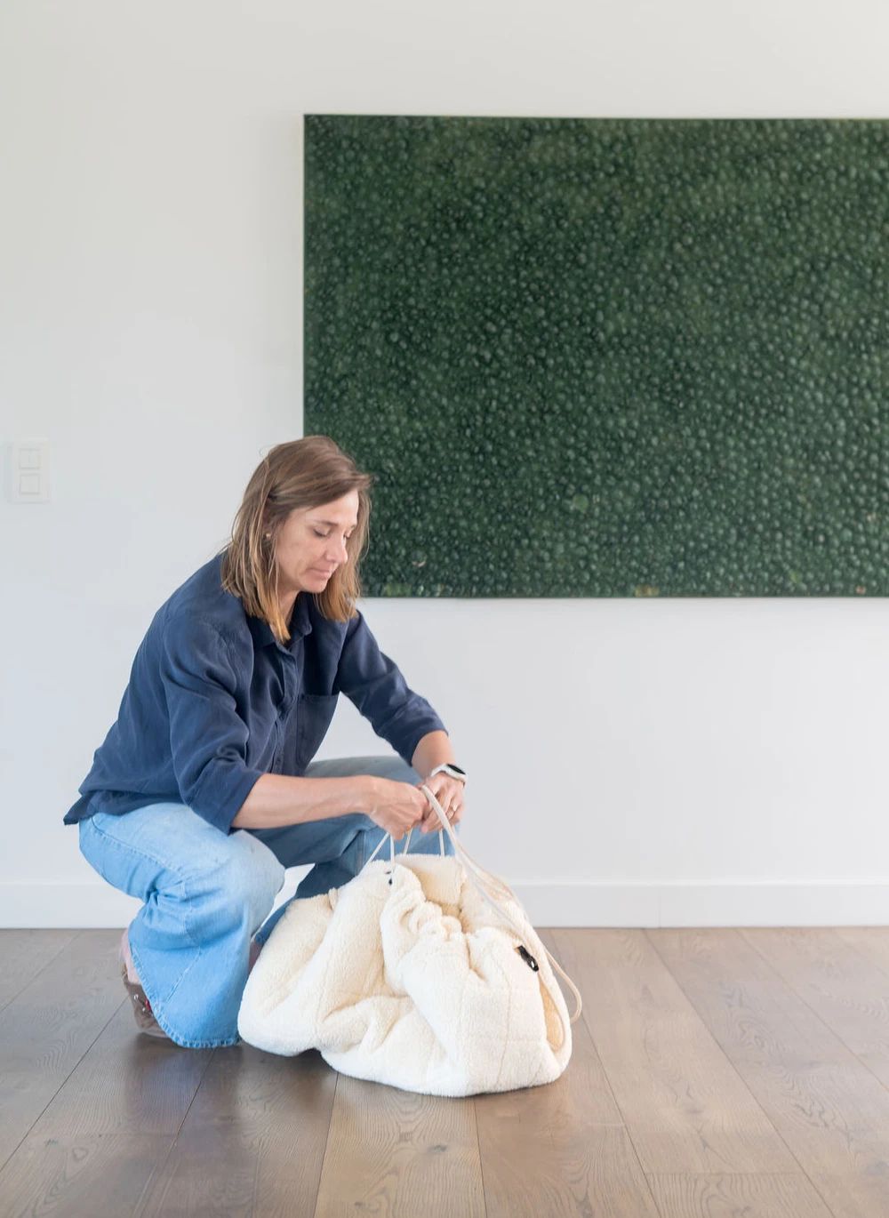A woman kneels on a wooden floor in a bright room, holding a large cream-colored plush drawstring bag. She is wearing a navy blue shirt and light blue jeans.