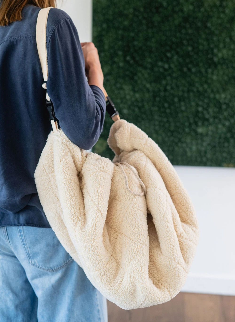 A person wearing a navy long-sleeve shirt and light blue jeans carries a large, soft cream-colored sherpa or fleece shoulder bag. The background features a white wall and a green textured wall panel.