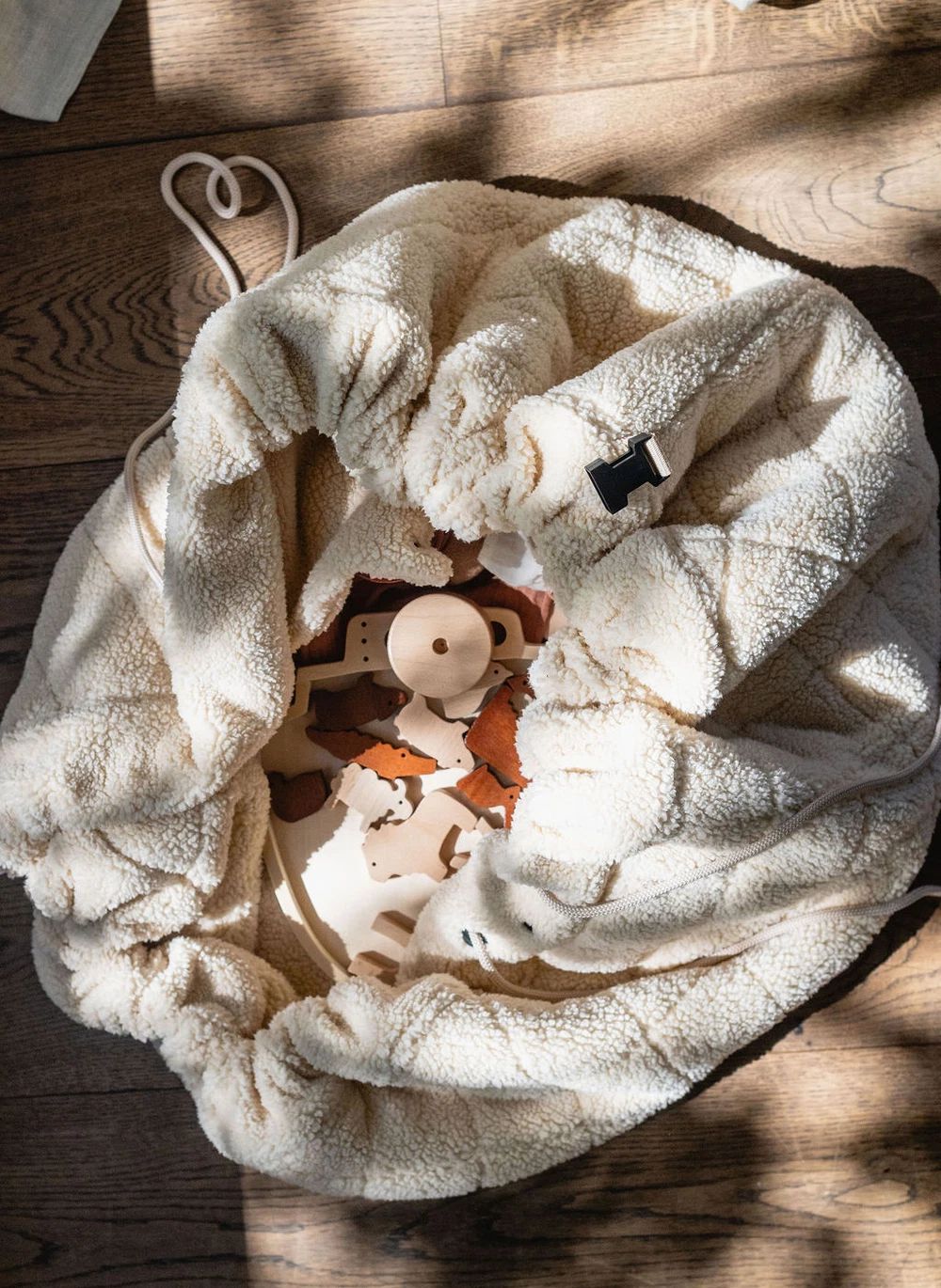 A cream-colored plush fleece drawstring bag filled with wooden toy pieces, photographed from above on a rustic wooden floor with dappled sunlight and shadows.