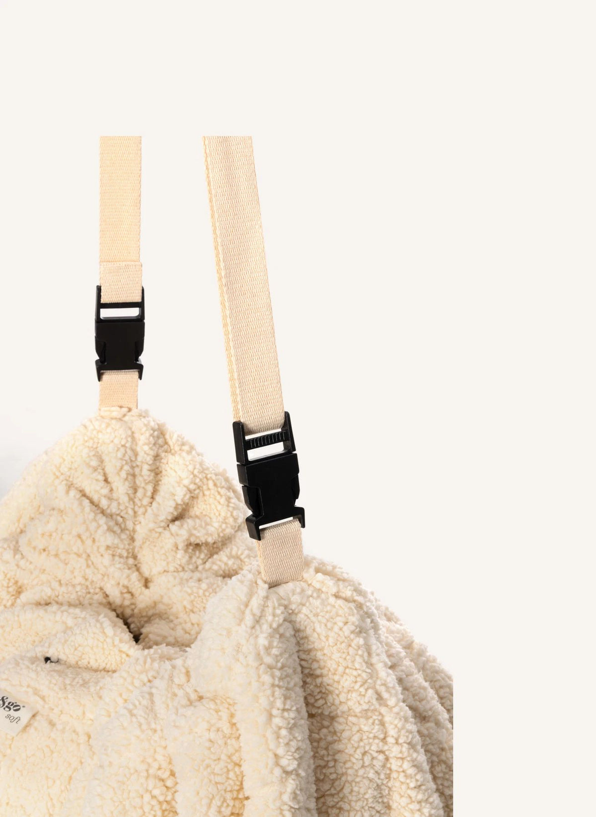 Close-up of a cream-colored sherpa or fleece drawstring bag with adjustable webbing straps and black plastic buckles, set against a plain off-white background.